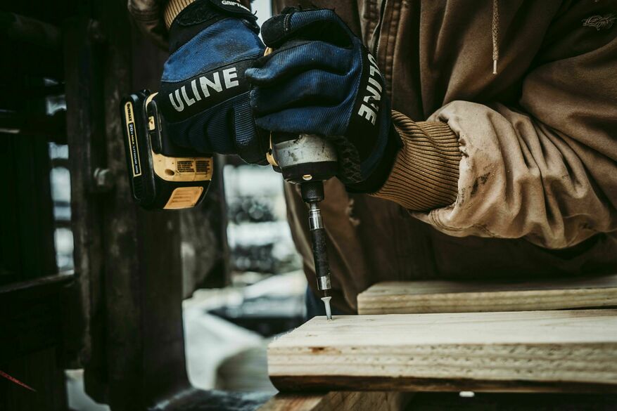 Close-up of a professional with protective blue gloves drilling a nail into a deck board.